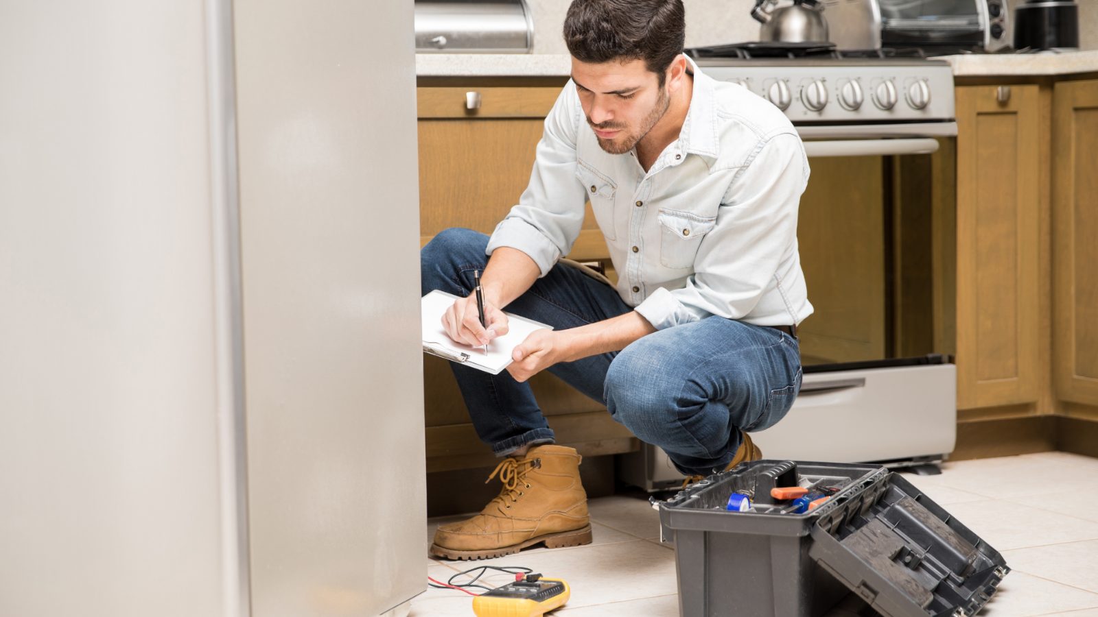 A contractor crouching in a kitchen to write on a clipboard, with an open toolbox and a multimeter on the floor beside him