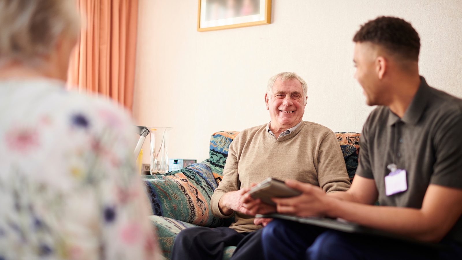 A support worker wearing an ID badge sitting with an older man and woman in their living room, looking at a tablet together